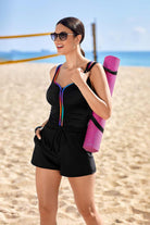 Woman on beach wearing a black swim romper with rainbow piping down the front and on the straps